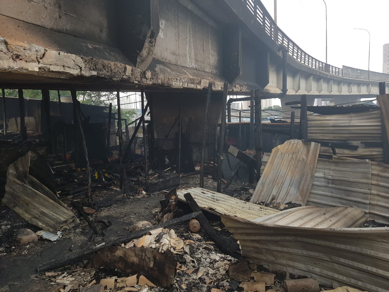 Imagem mostra área incendiada, entre os destroços, imagem da parte debaixo do viaduto com ferragens, pedaços de madeiras, tocos, pedras, telhas